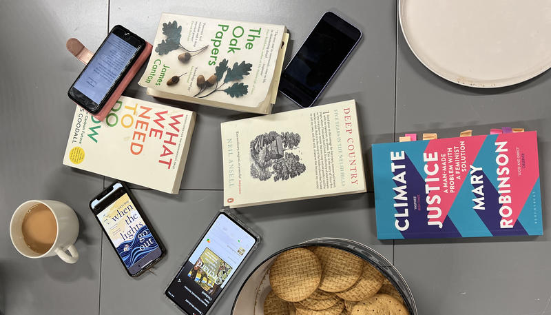Books and phones are scattered across a grey kitchen table, next to a biscuit tin and a cup of tea