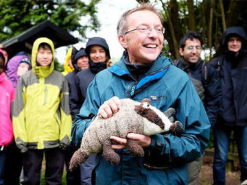 Nigel Fisher holds a plush toy badger, smiling while surrounded by people in brightly coloured rain coats