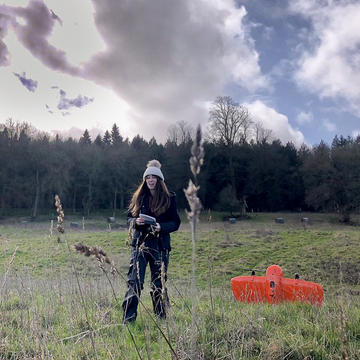 A female researcher stands in a field beside an orange drone, preparing to start a flight. She is wearing a bobble hat.